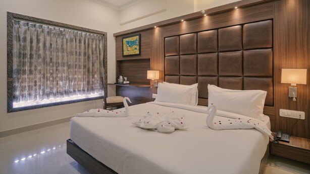 Habitación de hotel con una cama grande, cabecera acolchada, mesita de noche con lámpara y ventana con cortinas.