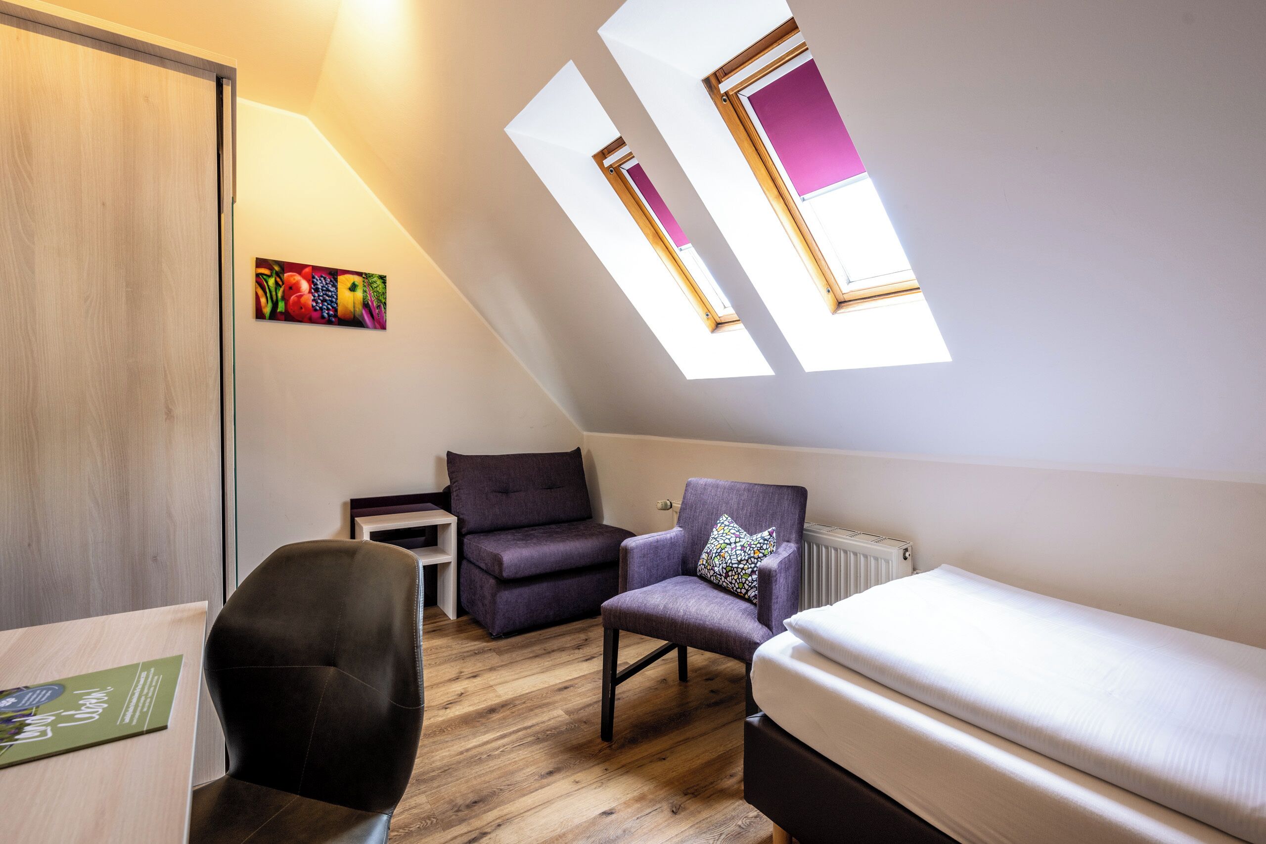 Una habitación pequeña y moderna con techo inclinado, cama, escritorio y dos sillas. Cuenta con tragaluces que permiten la entrada de luz natural.
