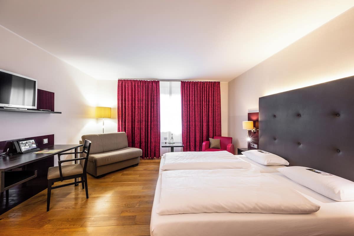 Une chambre d’hôtel avec un lit, un bureau équipé d’un ordinateur, une chaise, un canapé et un téléviseur fixé au mur.