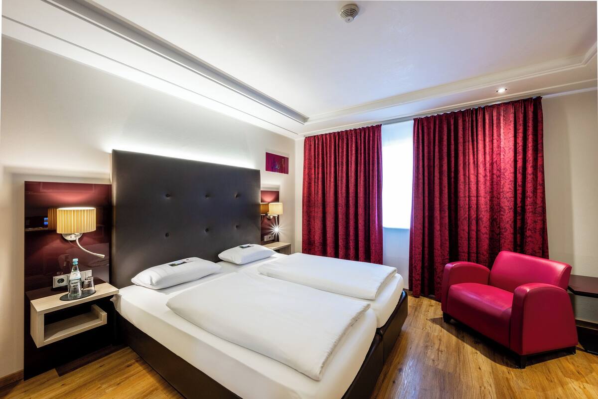 Une chambre d’hôtel avec un grand lit, un fauteuil rouge, une table de chevet avec une lampe et une fenêtre avec des rideaux rouges.