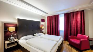 Una habitación de hotel con una cama grande, un sillón rojo, una mesita de noche con una lámpara y una ventana con cortinas rojas.