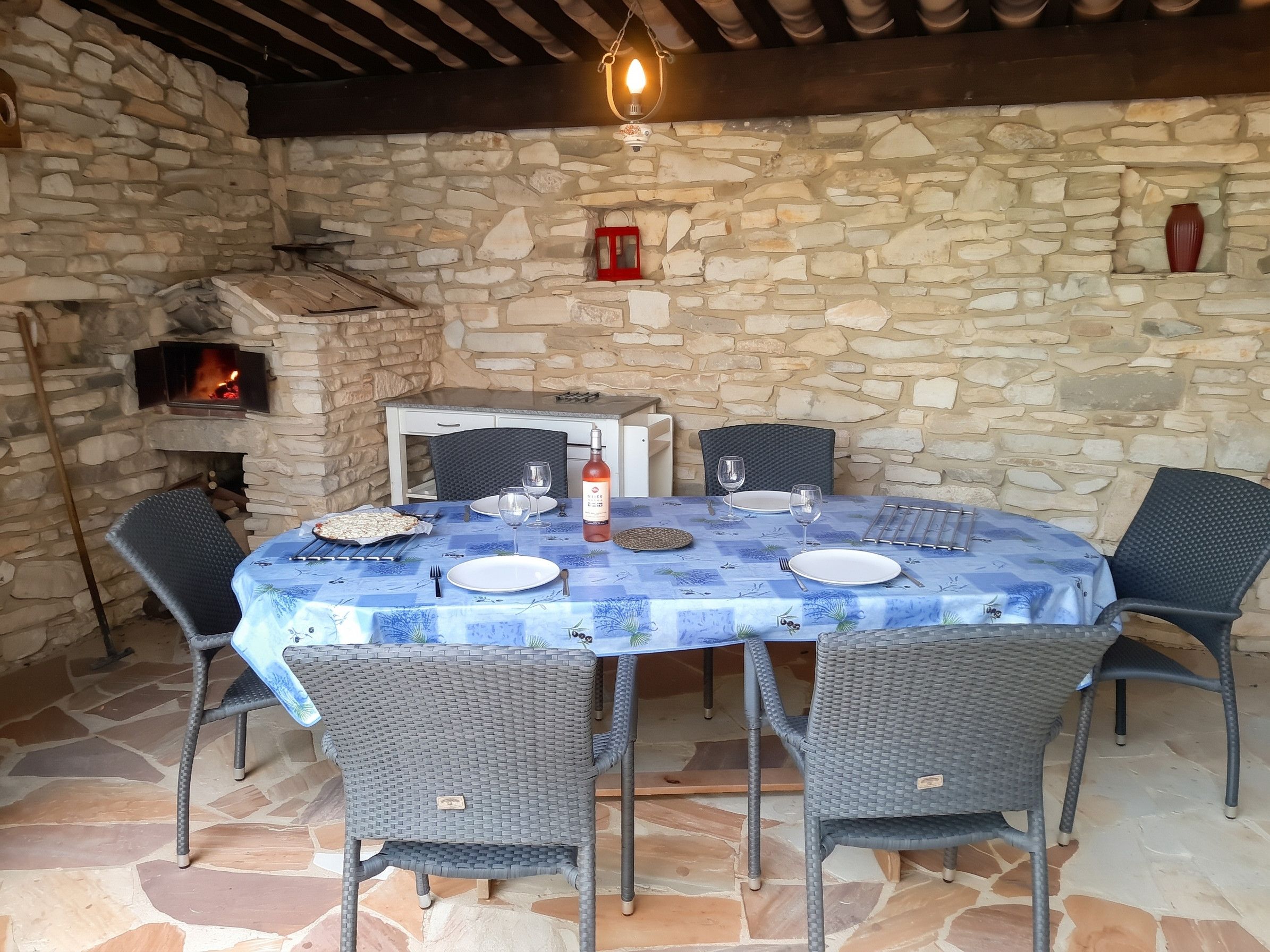 Un espace extérieur aménagé pour les repas, avec des murs en pierre, une table dressée pour deux personnes et entouré de chaises en osier.