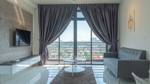 A modern living room with a grey sofa, a flat-screen TV mounted on the wall, and a large window with curtains.