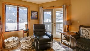 A cozy living room with a leather chair, a wooden table, and a couch. Large windows offer a view of snow-covered mountains. - Walk to Copper Mountain Lifts: Mtn-View Condo (Copper Mountain)
