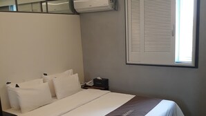 A hotel room with a bed, a nightstand, a telephone, a window with blinds, and an air conditioning unit.