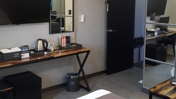 A modern hotel room with a wooden desk, a flat-screen TV mounted on the wall, a large mirror, and a bed with a branded bedspread.