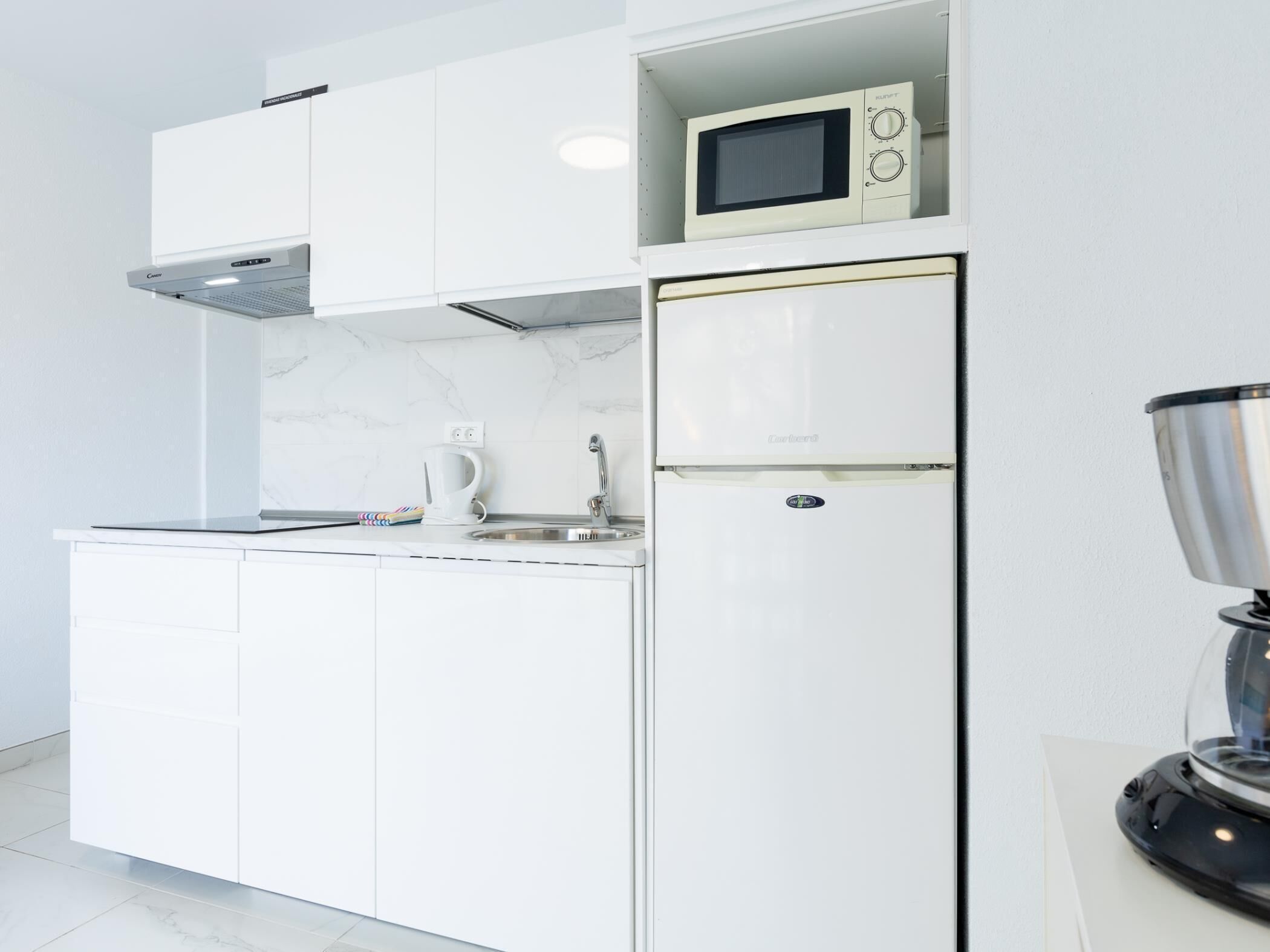 A modern kitchen with white cabinetry, a built-in microwave, a stainless steel sink, and a coffee maker.