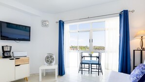 Una habitación de hotel moderna con una televisión de pantalla plana, cafetera, ventilador, mesa de comedor con sillas y un amplio ventanal con cortinas.