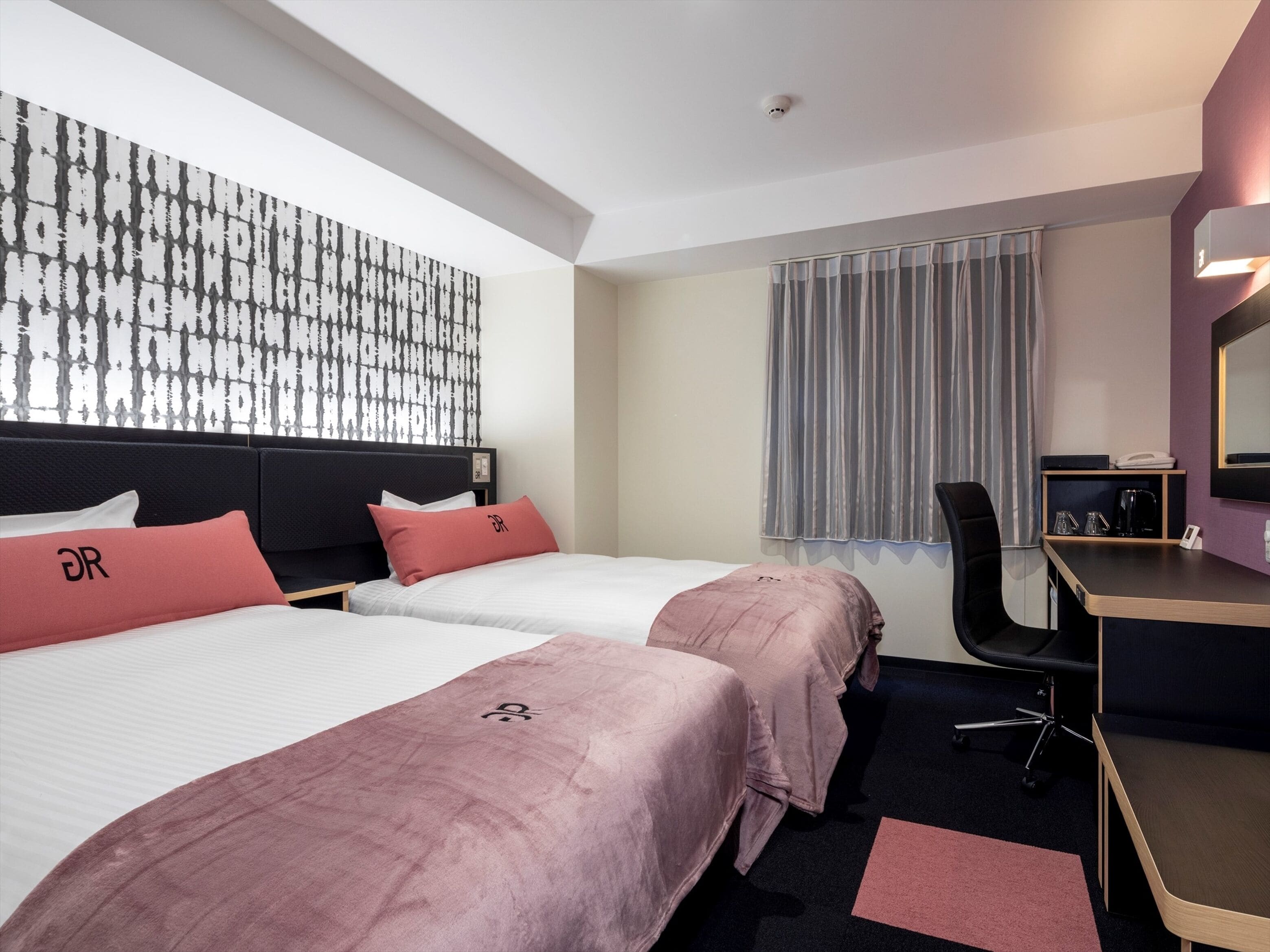 Habitación de hotel con dos camas, un escritorio, una silla, un ventanal con cortinas y un espejo montado en la pared.