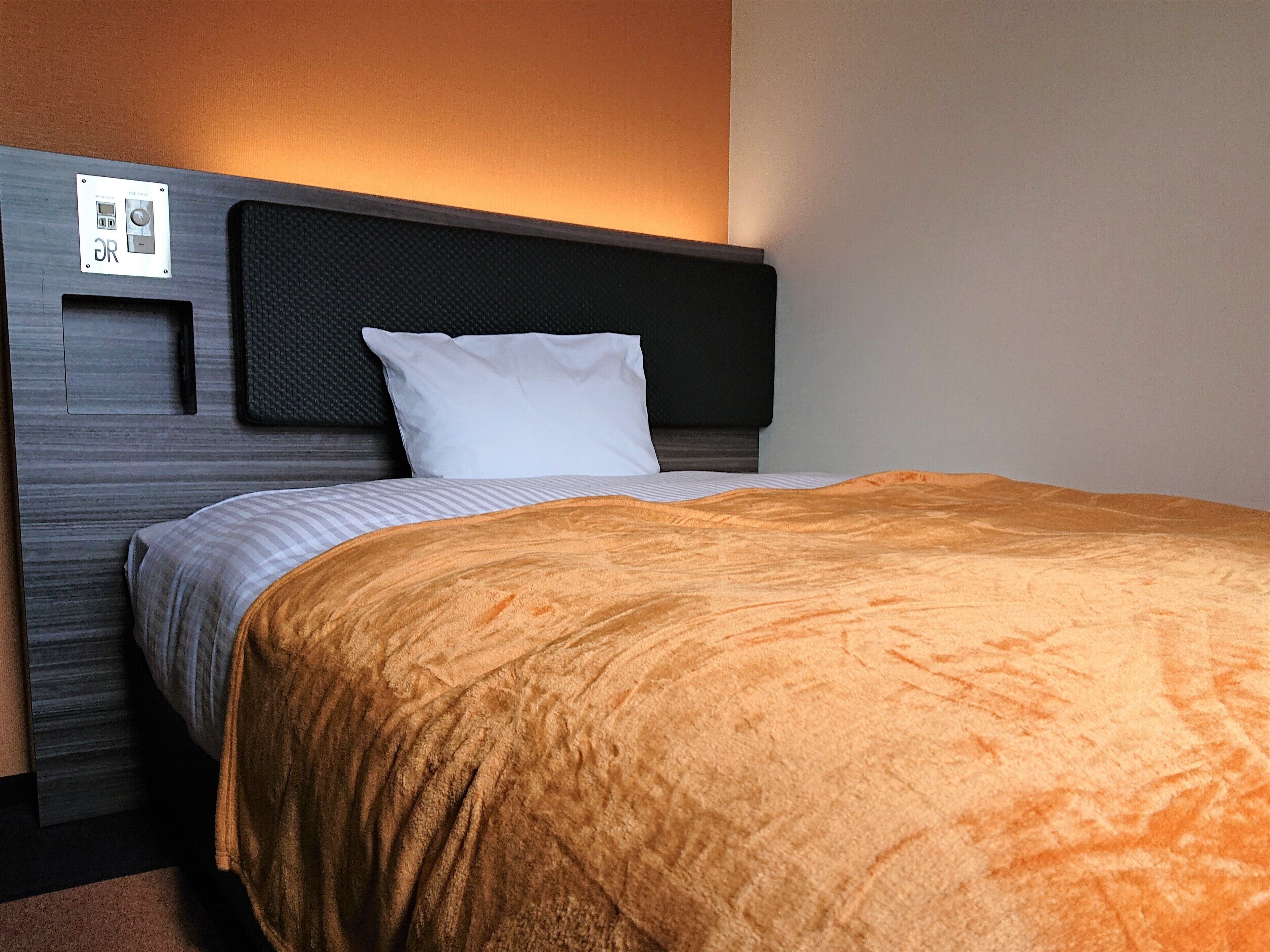 A hotel room with a bed, a headboard, a control panel, and a wall-mounted light.