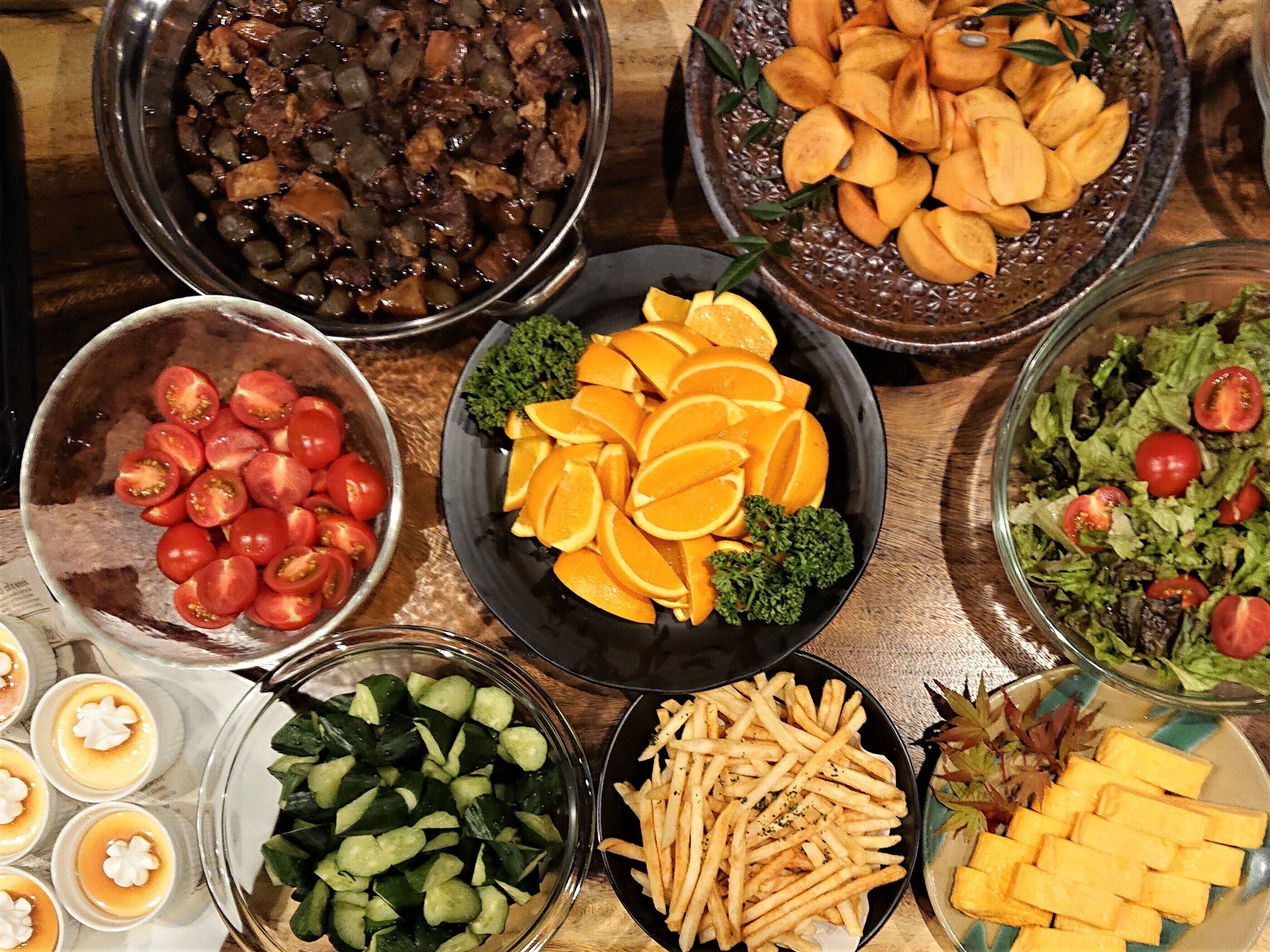 A variety of food items including a bowl of sliced tomatoes, a bowl of sliced oranges, a bowl of mixed vegetables, a bowl of fries, and a bowl of salad.