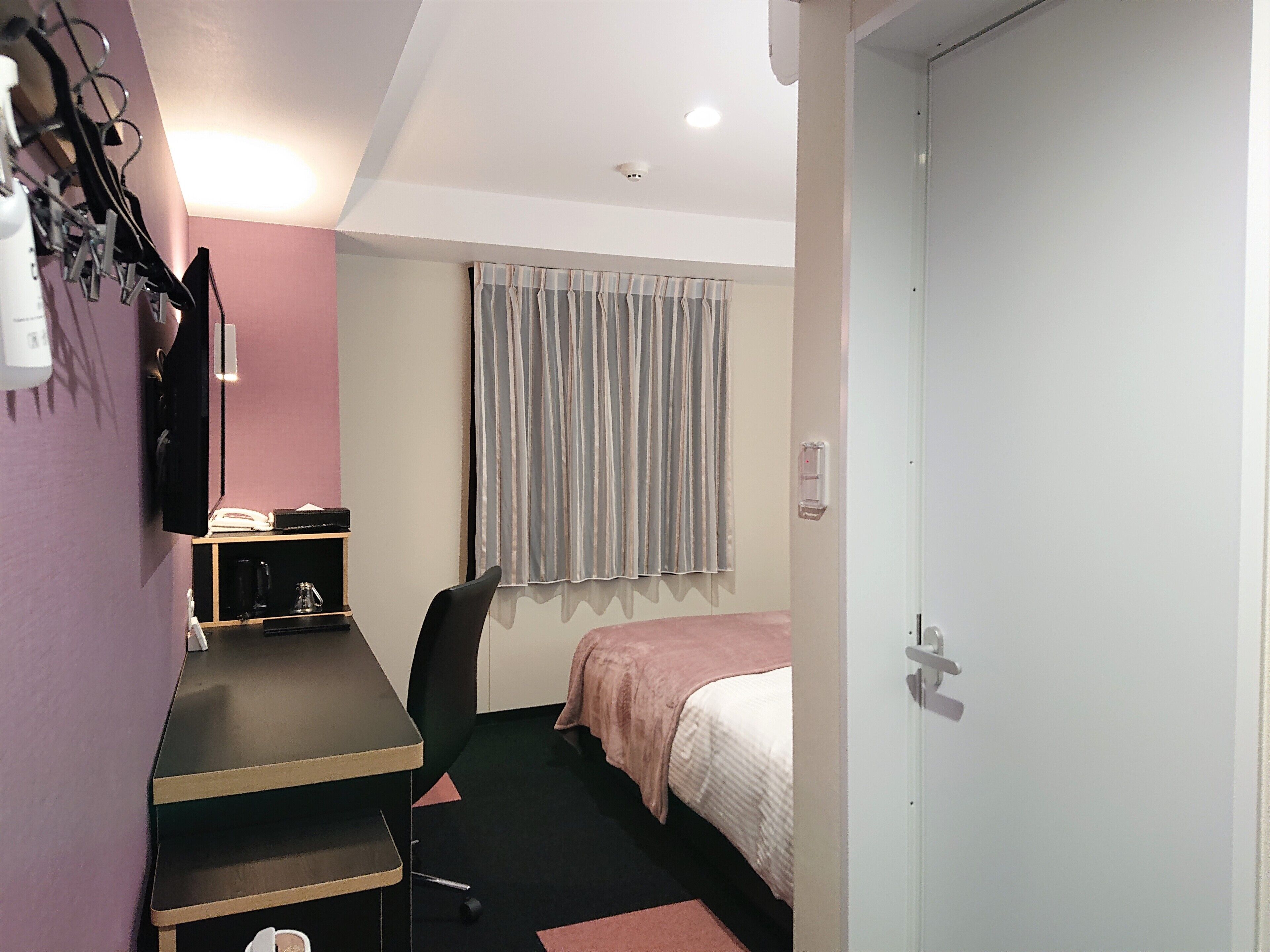Une chambre d’hôtel de taille réduite, comprenant un lit, un bureau avec une chaise, une petite table, un téléphone et une fenêtre avec des rideaux.
