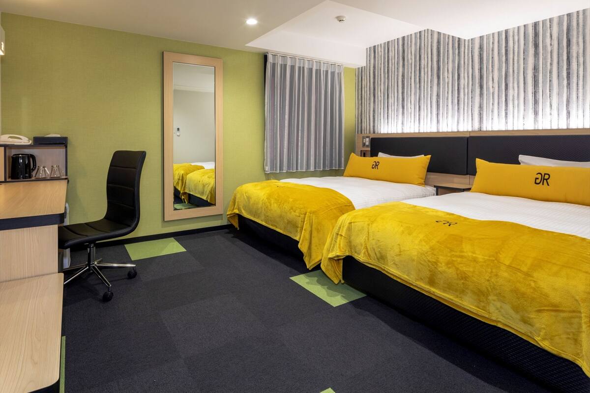 Ein Hotelzimmer mit zwei Betten, einem Schreibtisch, einem Stuhl, einem Spiegel und einem Fenster mit Vorhängen.
