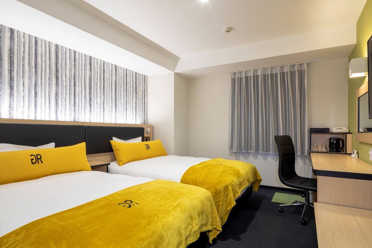 Ein Hotelzimmer mit zwei Betten, einem Schreibtisch, einem Stuhl, einer Kaffeemaschine und einem Fenster mit Vorhängen.