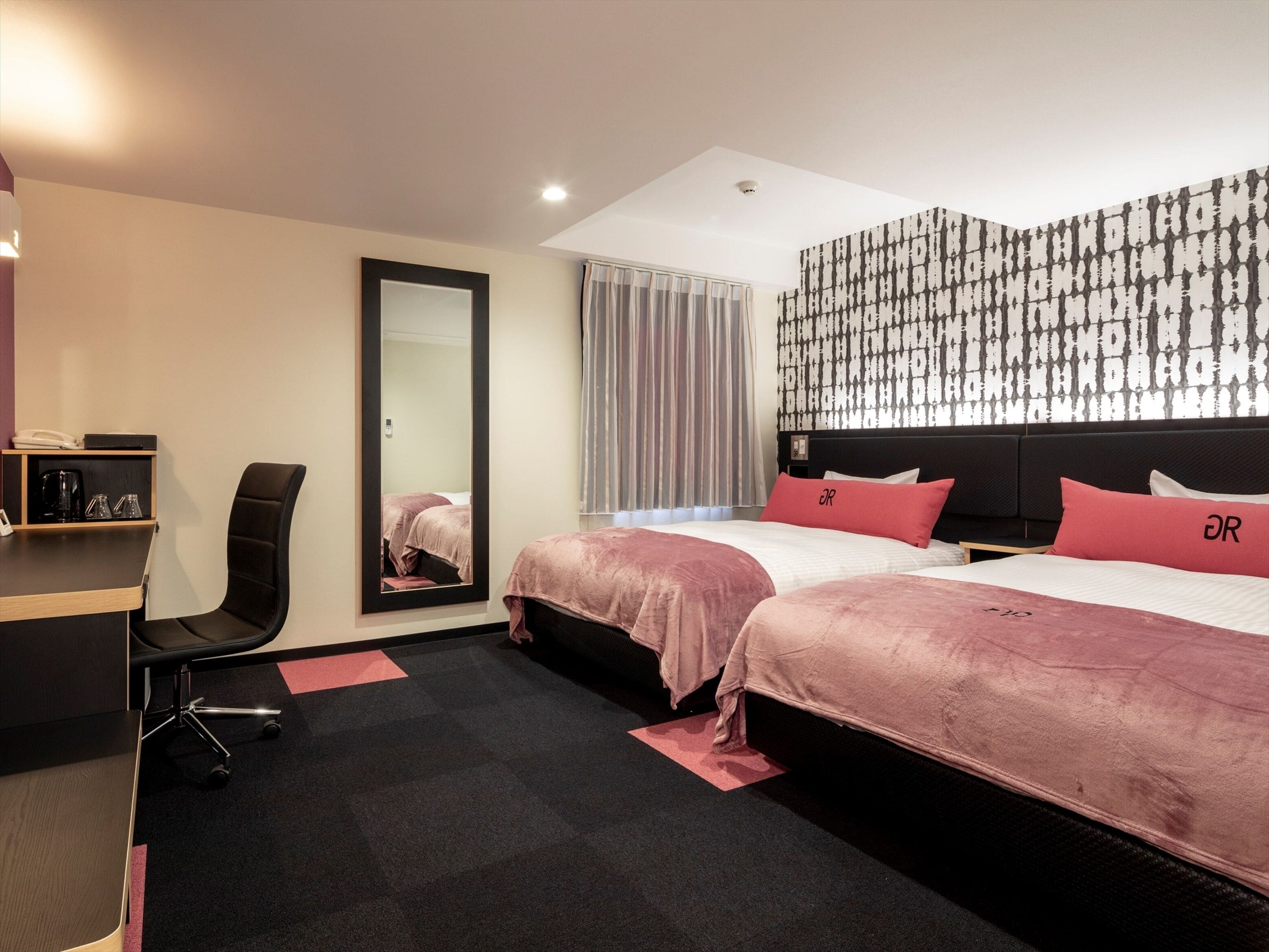 A hotel room with two beds, a desk, a chair, a large mirror, and a patterned wall.