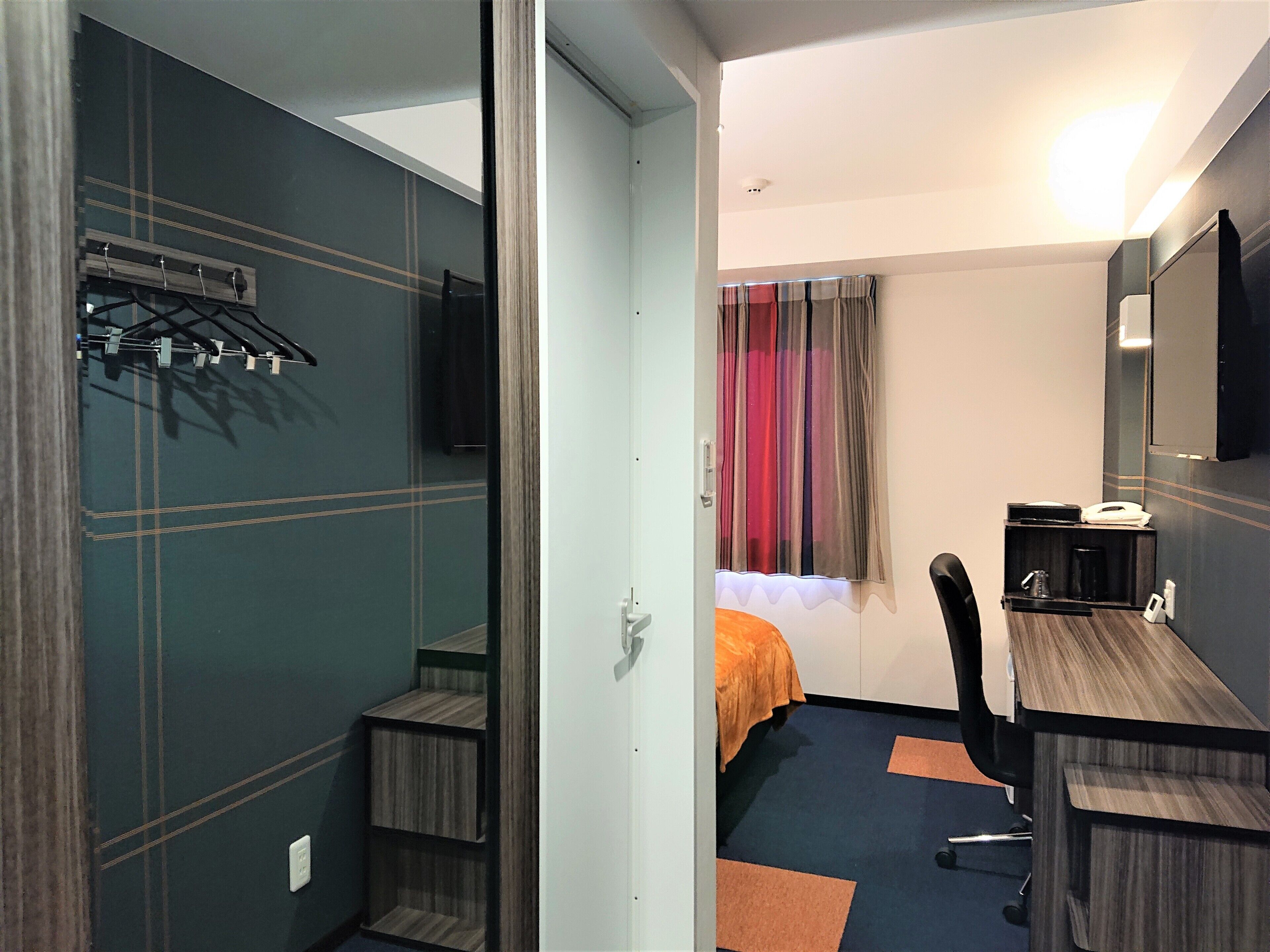 A hotel room with a bed, a desk, a chair, a TV, and a coat rack.