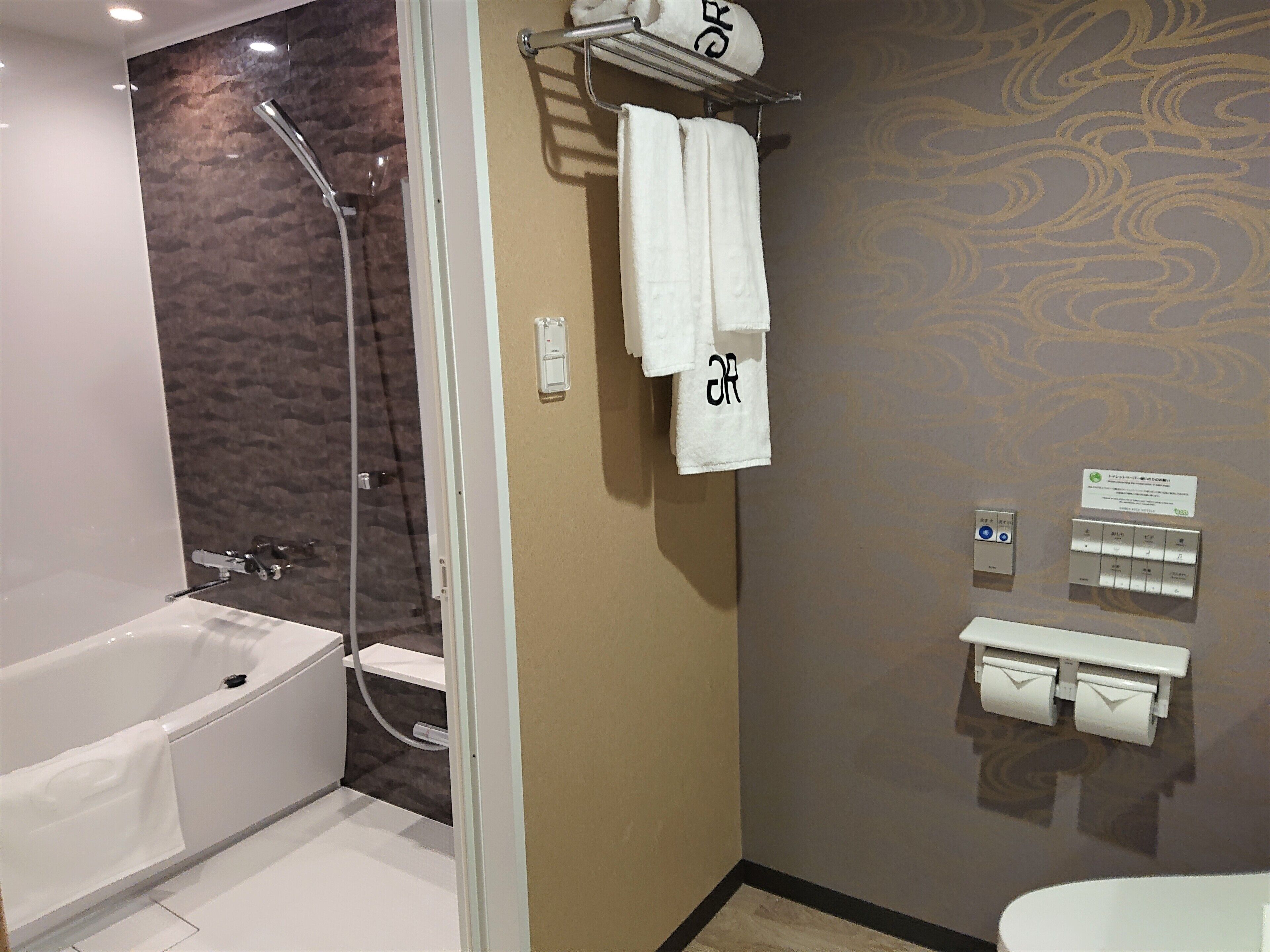 A modern bathroom with a bathtub, shower, and toilet. There are towels hanging on a rack and a paper towel dispenser.