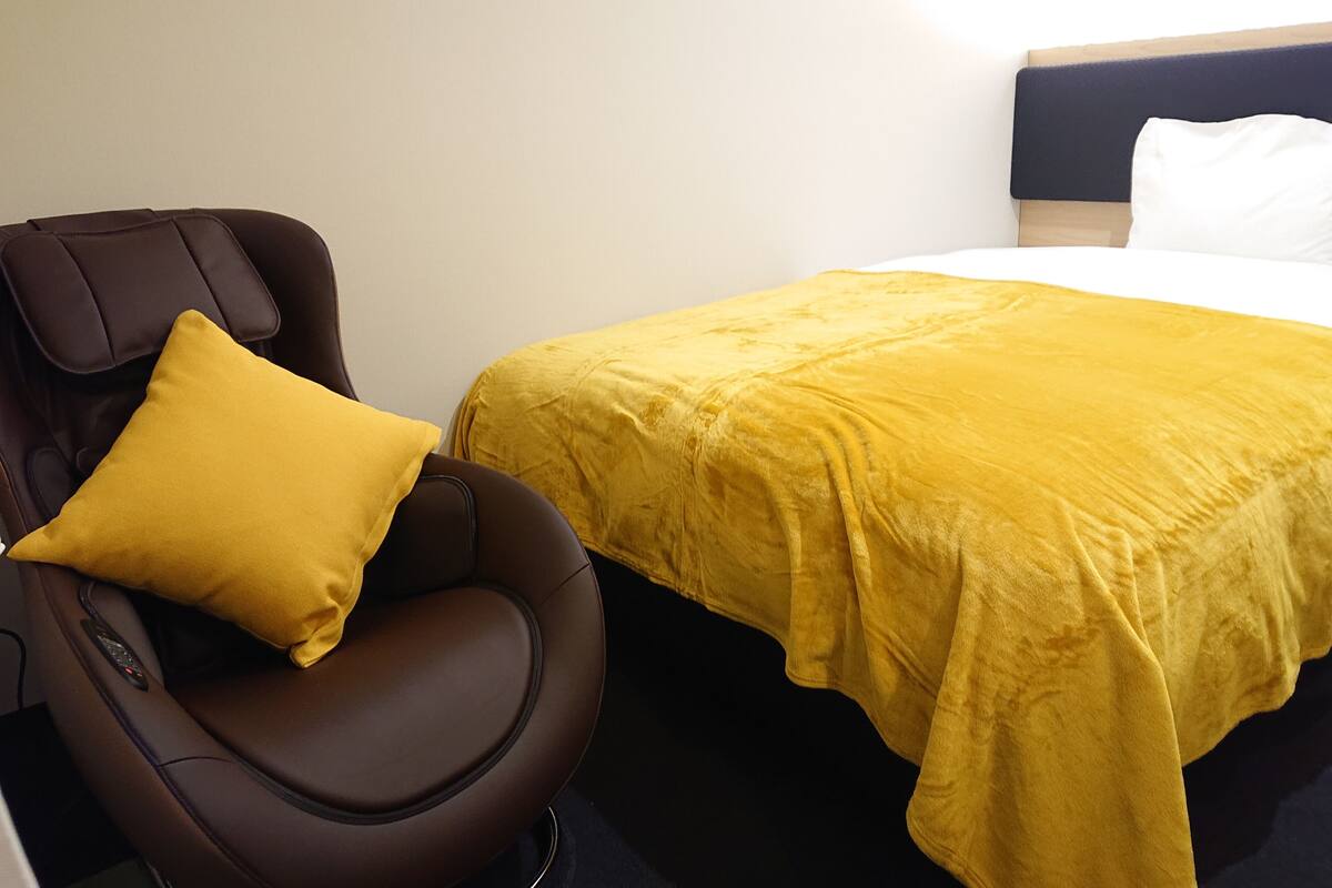Ein Zimmer mit Massagesessel, einem Bett mit gelber Bettdecke und einem weißen Kopfkissen.