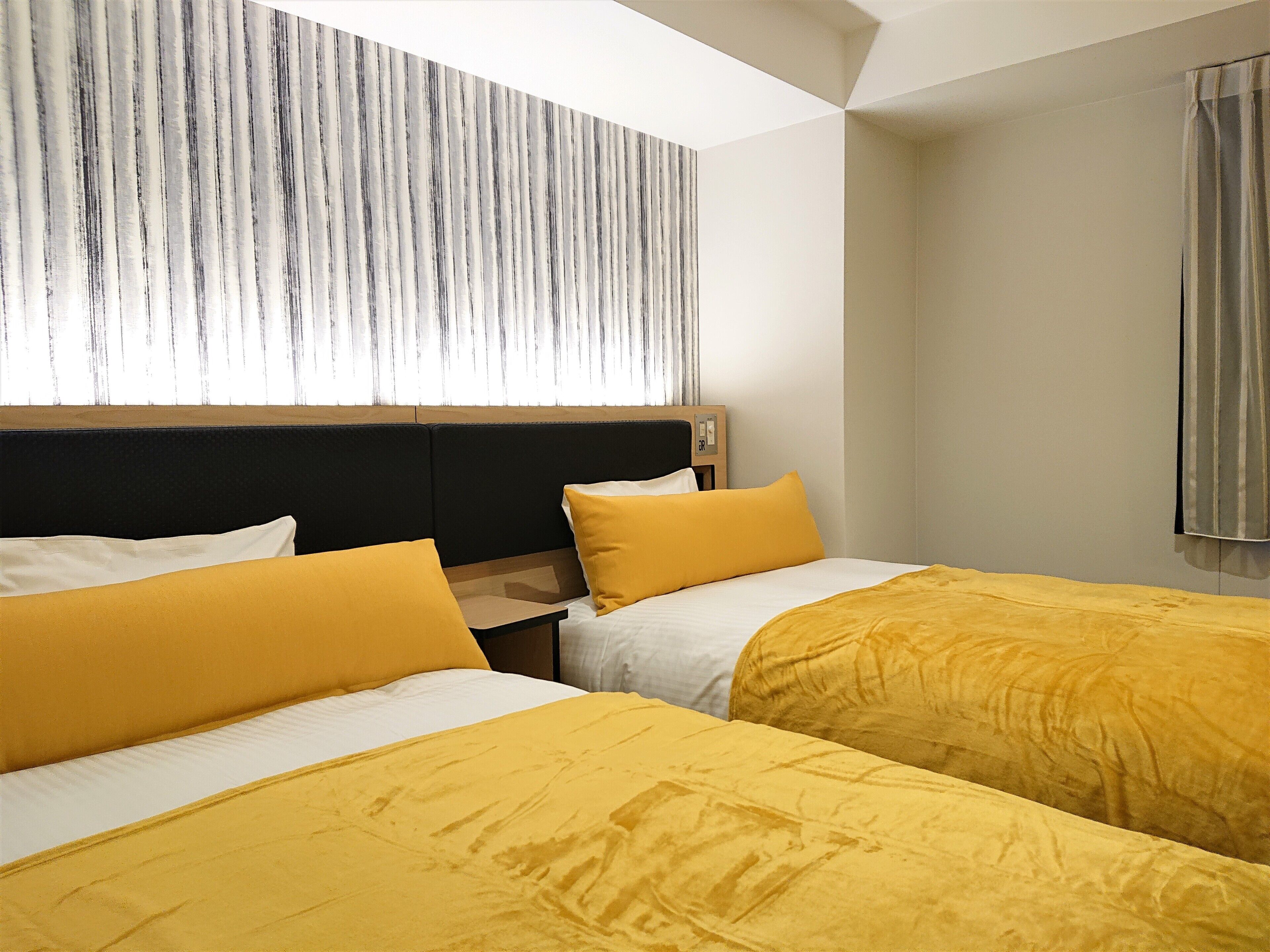 A hotel room with two beds, each with yellow bedding, a wooden headboard, and a window with sheer curtains.