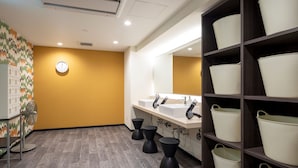 Ein modernes Badezimmer mit Holzboden, einer gelben Akzentwand und einem großen Spiegel über den Waschbecken. Schwarze Hocker und ein Ablageschrank mit weißen Behältern sind ebenfalls vorhanden.