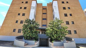 Ein mehrgeschossiges Gebäude mit einer symmetrischen Fassade, wobei ein zentraler Eingang von zwei großen Pflanzkübeln mit Bäumen eingerahmt wird.