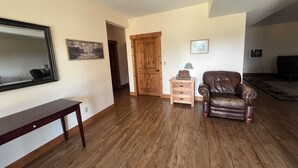 A living room with hardwood flooring, a brown leather armchair, a wooden side table with a lamp, a framed picture on the wall, and a mirror.