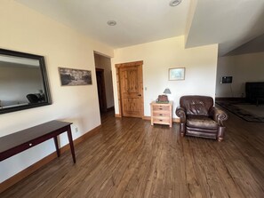 A living room with hardwood flooring, a brown leather armchair, a wooden side table with a lamp, a framed picture on the wall, and a mirror.