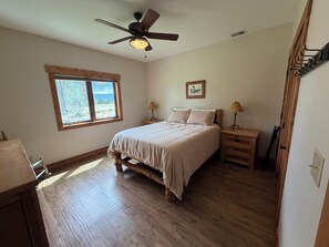 A bedroom with a wooden floor, a bed with a headboard, a ceiling fan, a window with a view, and a nightstand with a lamp.