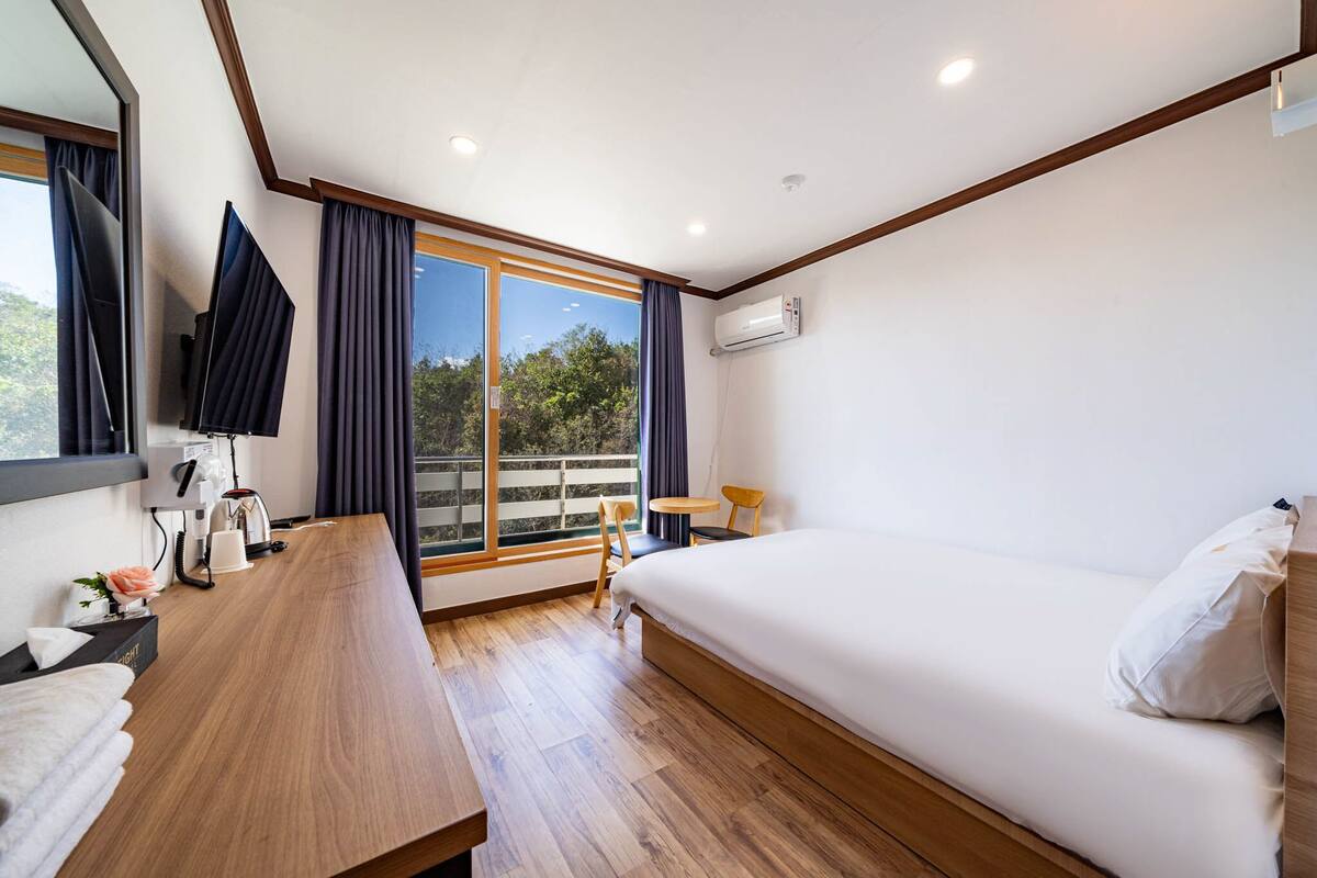 A modern hotel room with a wooden desk, a bed, a TV, and a balcony with curtains.