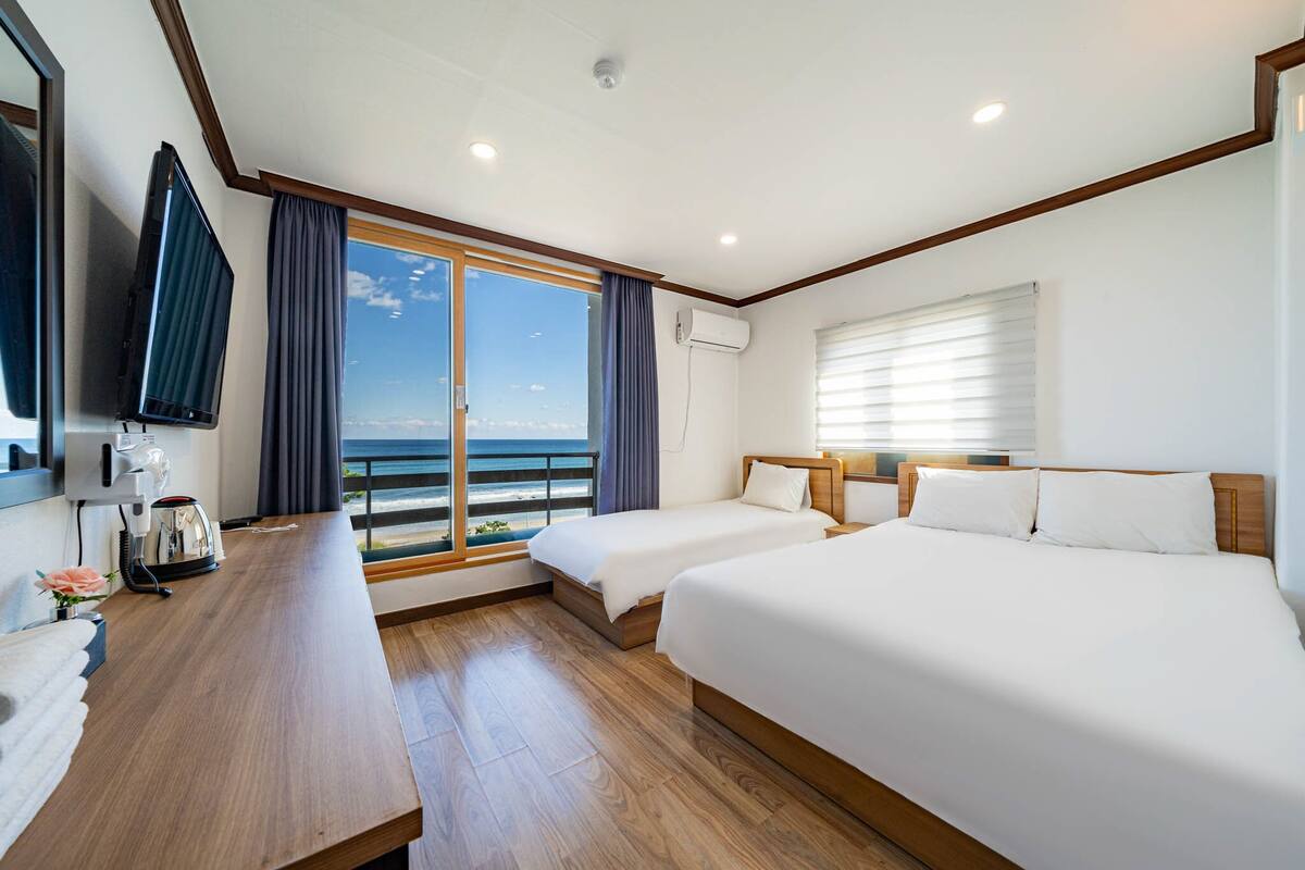 A hotel room with two beds, a wooden desk, a TV, and a view of the ocean.
