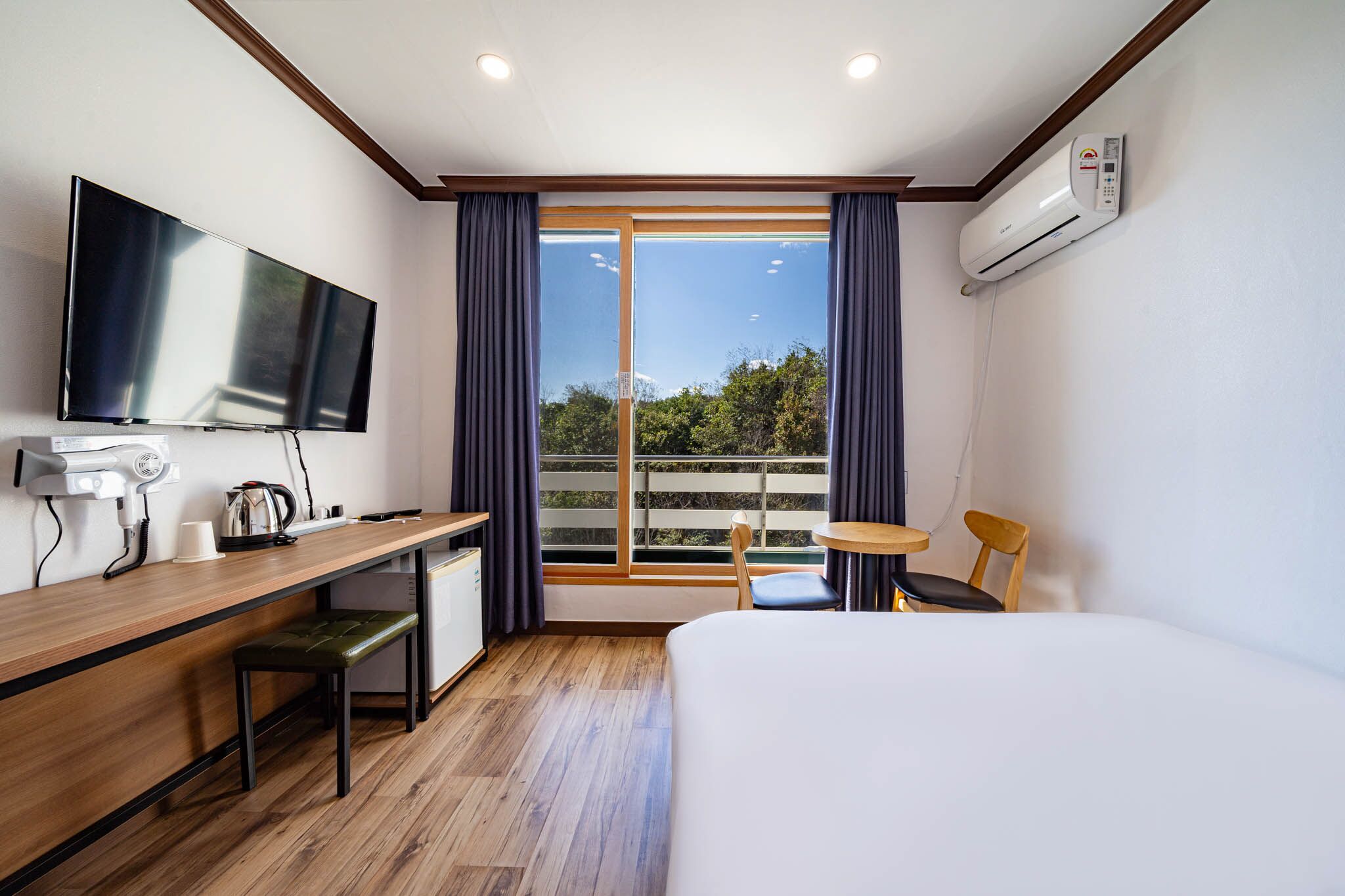 A hotel room with a bed, a desk with a TV, a coffee maker, a chair, and a view of greenery through large windows.