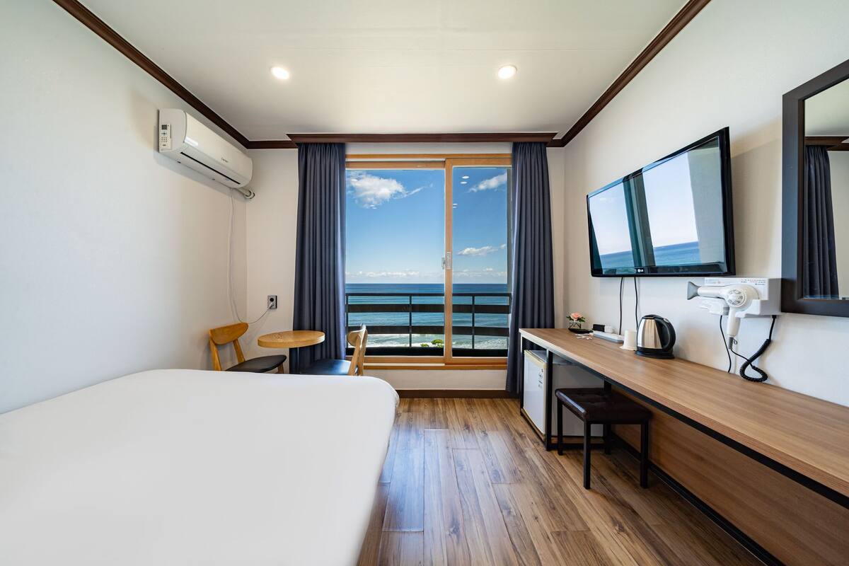 A hotel room with a bed, a desk, a chair, a small table, a TV, and a view of the ocean.