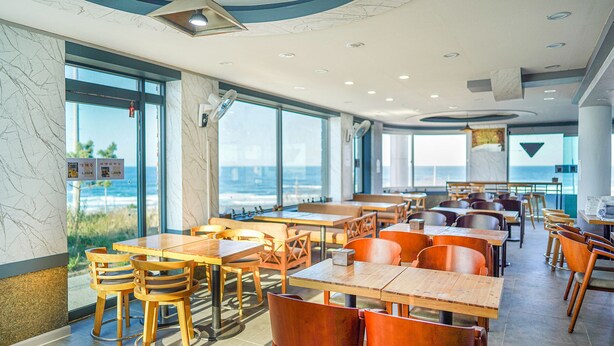A spacious dining area with large windows offering a view of the sea, wooden tables and chairs, and a modern ceiling design. - Mangsang Beach Pension (Gangneung)