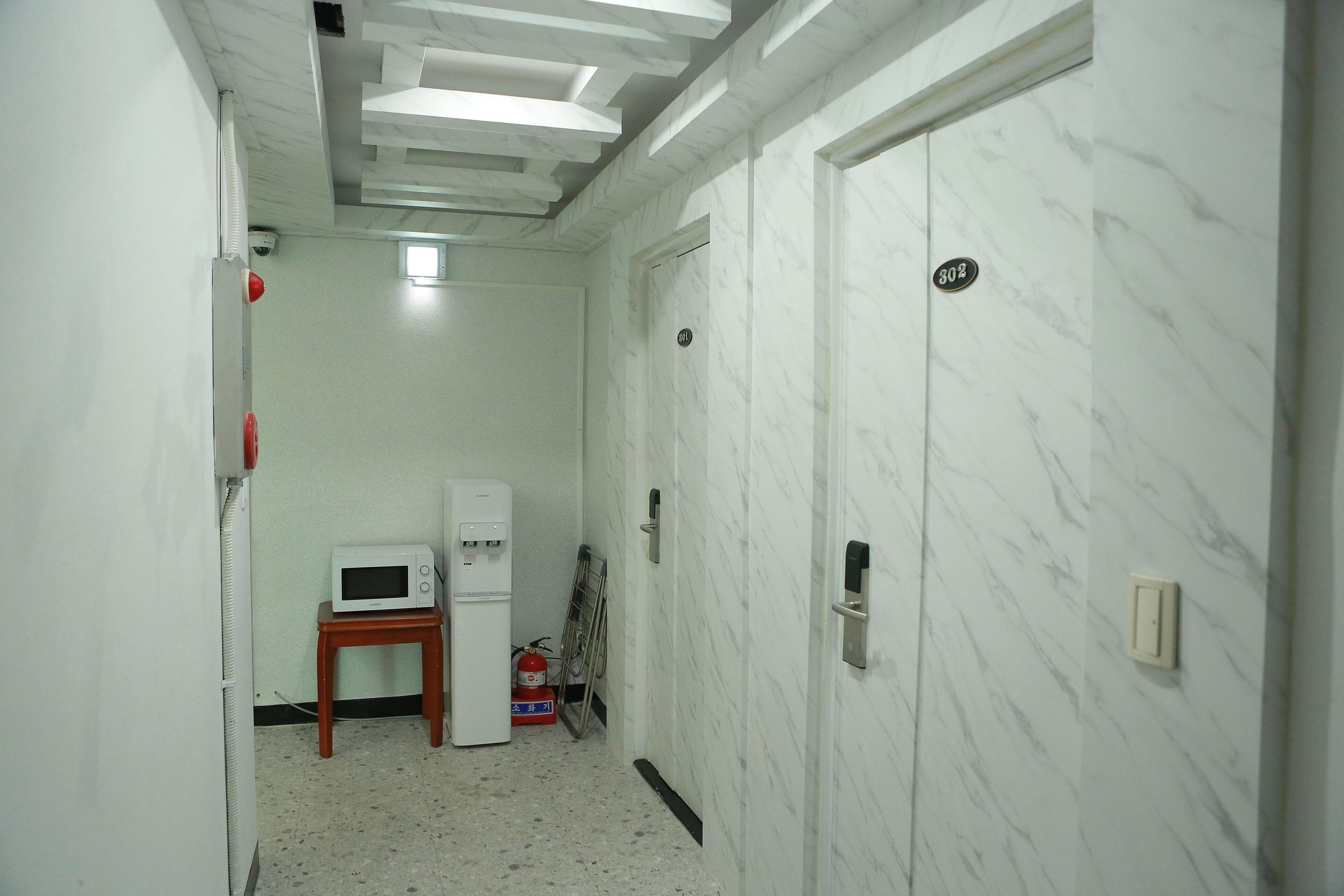 A narrow hallway with white marble walls, a white door with a number plate, a small table with a microwave, and a fire extinguisher.