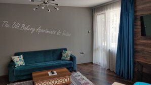 A modern living room with a teal sofa, a wooden coffee table, and a patterned rug. A ceiling light fixture is visible, and the wall has a sign reading "The Old Bakery Apartments & Suites". - The Old Bakery Apartments (Plovdiv)