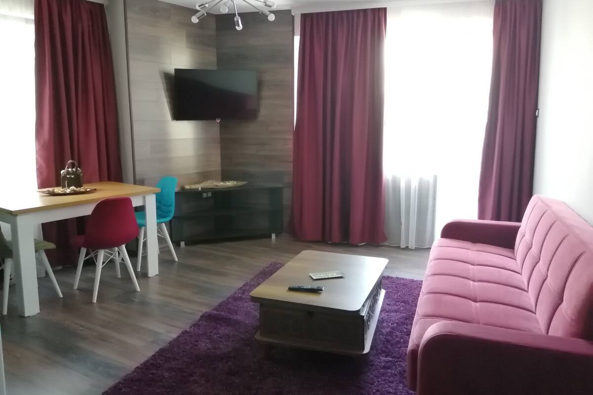 A modern living room with a sofa, a coffee table, a TV mounted on the wall, and a dining area with a table and chairs.