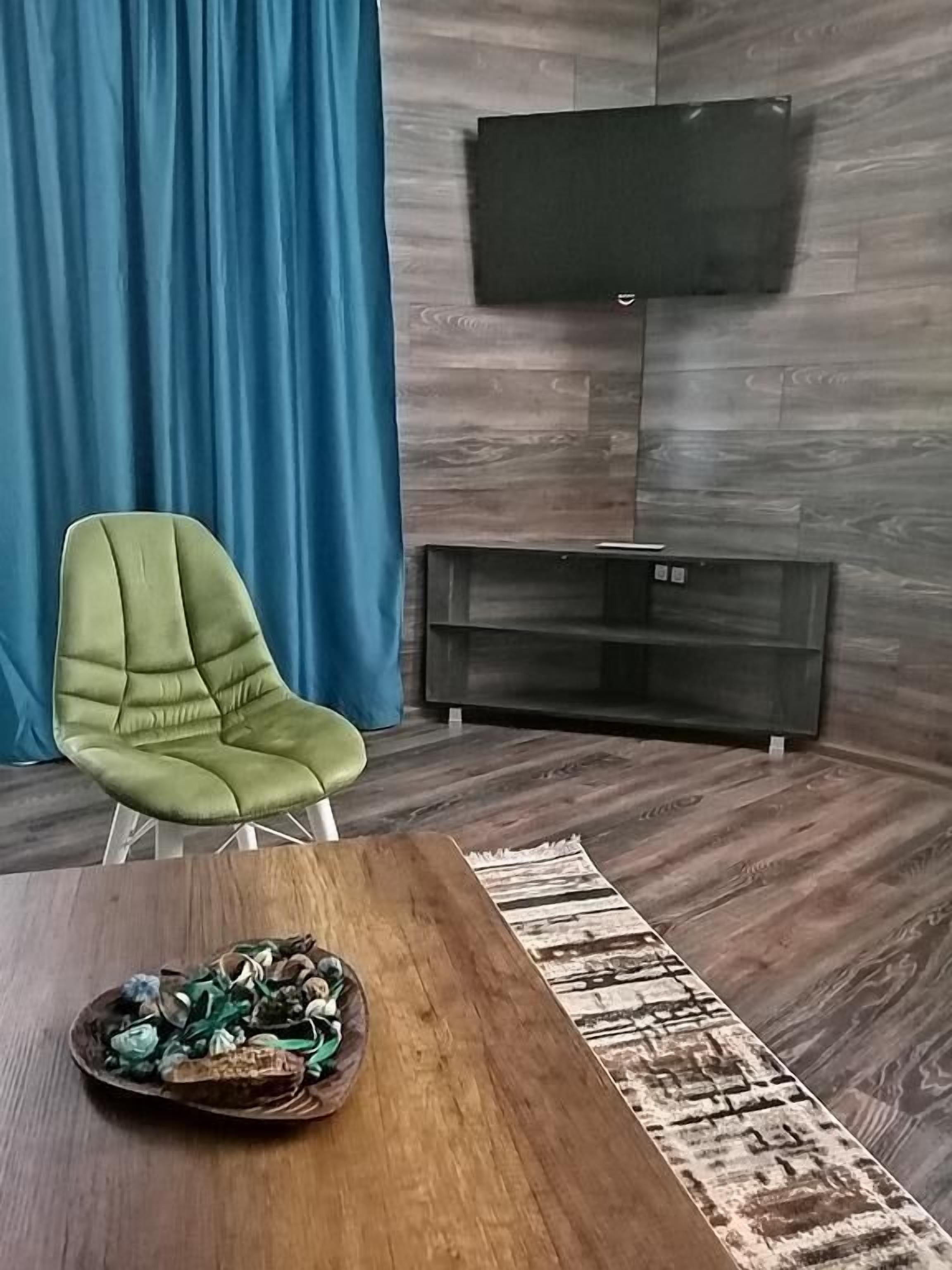 Ein modernes Wohnzimmer mit einem grünen Sessel, einem Holztisch, einem an einer Holzwand montierten Flachbildfernseher und einem blauen Vorhang.