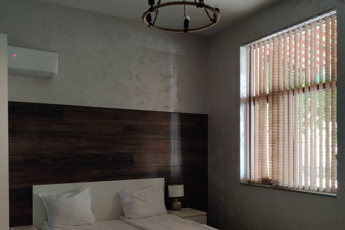 A modern bedroom with a wooden headboard, a bed with white linens, a bedside table with a lamp, and a window with vertical blinds.
