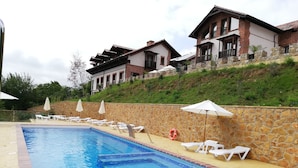 A swimming pool with lounge chairs and umbrellas, surrounded by stone walls and a hillside with buildings. - Hotel Cerro la Nina (Cangas de Onis)