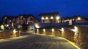 Une allée bordée de murs en pierre mène à une rangée de maisons dont les fenêtres sont illuminées, sous un ciel dégagé.