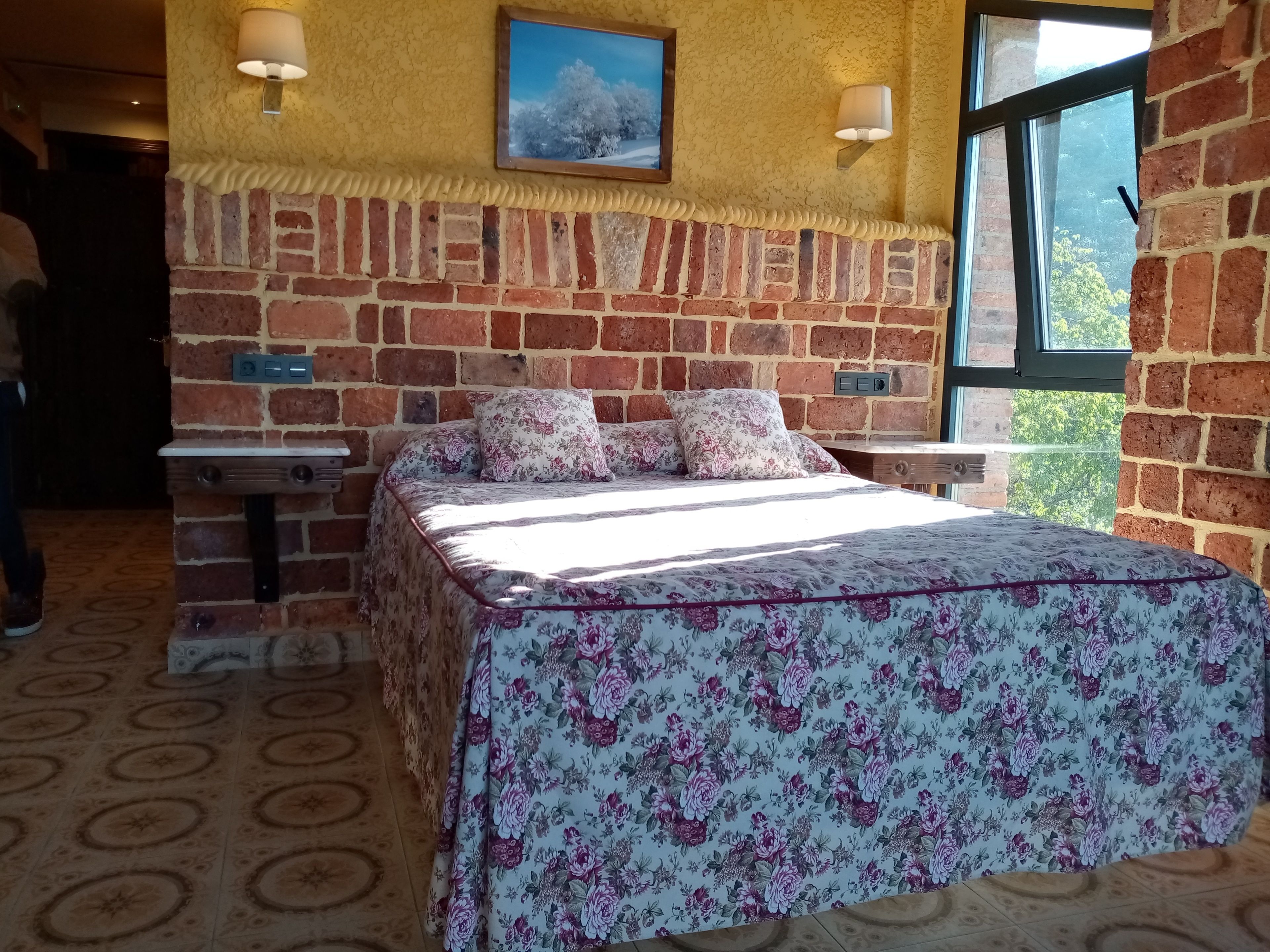 A room with a brick wall, a bed with floral bedding, two wall-mounted lamps, and a window with a view of trees.