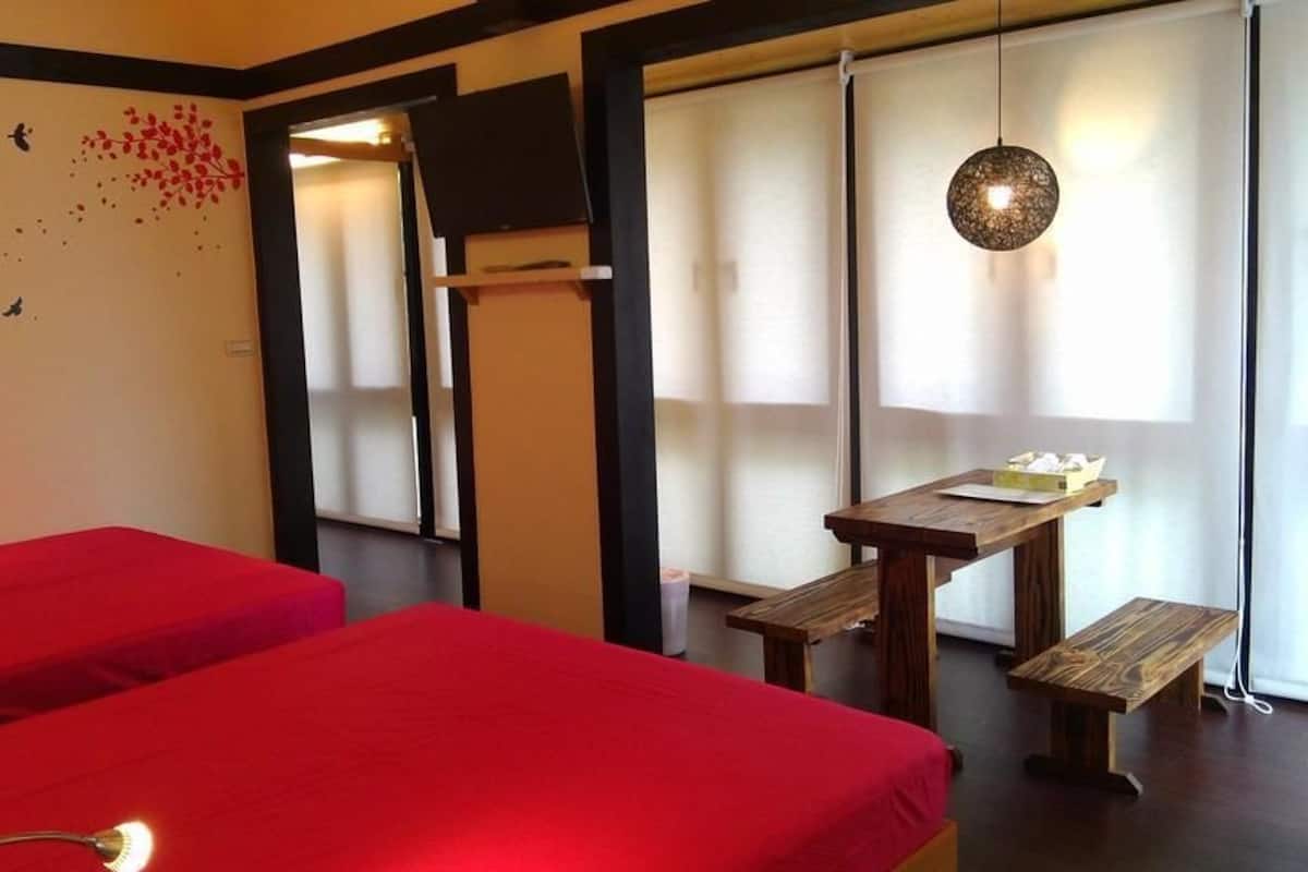 A room with two red beds, a wooden table with benches, a wall-mounted TV, and a hanging light fixture.