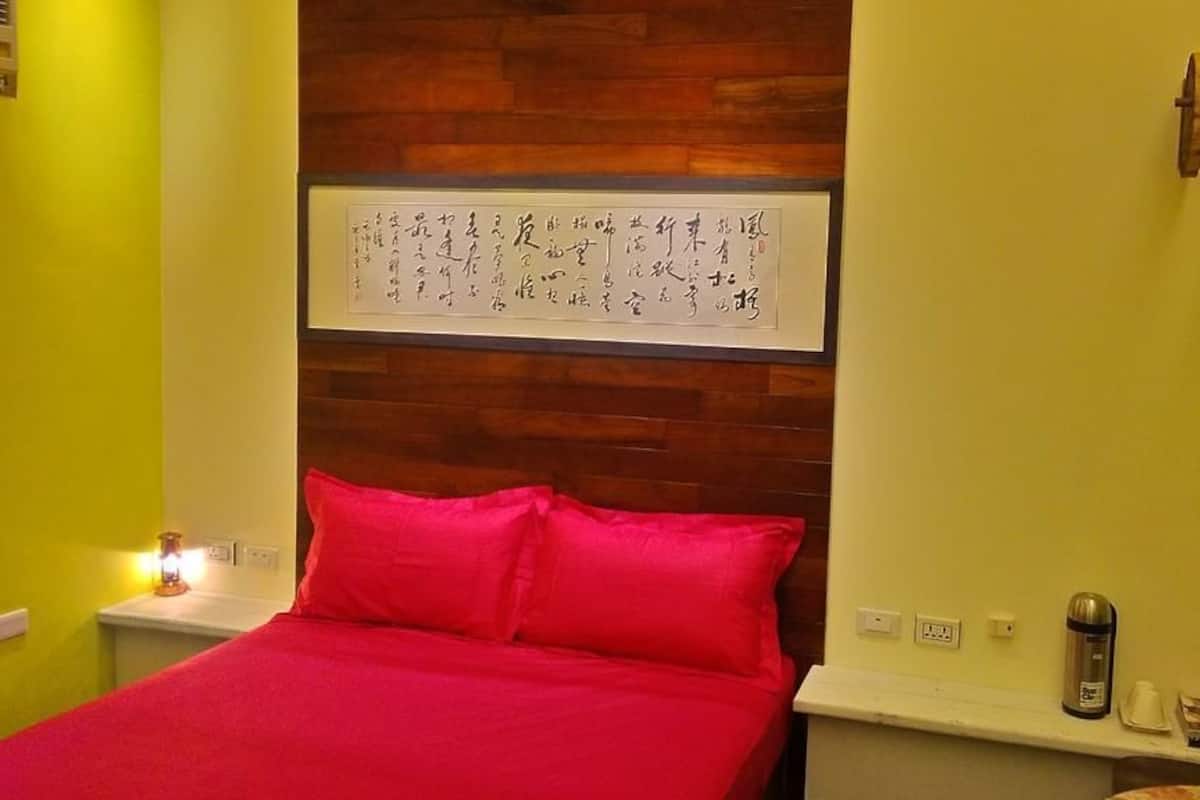 A room with a wooden headboard, a bed with red bedding, a wall-mounted artwork, a bedside table with a lamp, and a small shelf with items.