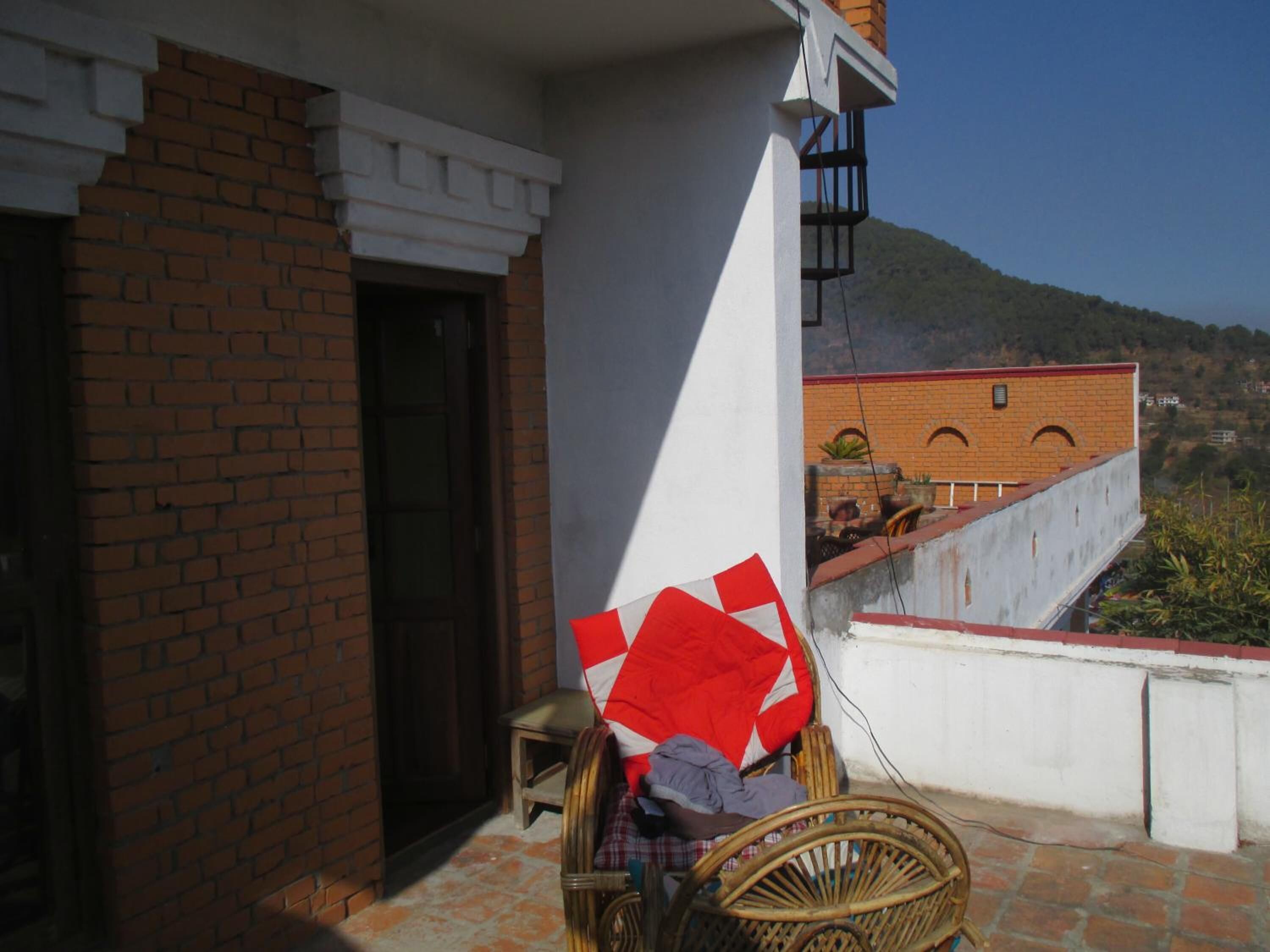 Un balcon avec un fauteuil en osier, un objet rouge et blanc, et une vue sur un bâtiment avec un toit rouge et une montagne en arrière-plan.