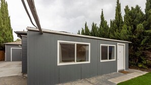 Un edificio de madera gris de una sola planta con dos ventanas y una puerta, rodeado de césped y árboles altos.