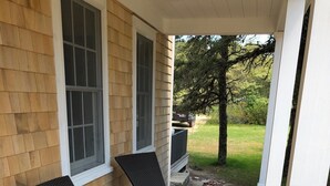 A covered porch with two wicker chairs and a small table, a white planter, and a view of a grassy yard. - Modern 5 Bedroom Home. Newly constructed. Large yard. Close to beaches and town. (Nantucket)