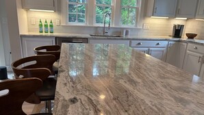A kitchen with a large marble island, wooden chairs, and a view of trees through the windows. - Modern 5 Bedroom Home. Newly constructed. Large yard. Close to beaches and town. (Nantucket)