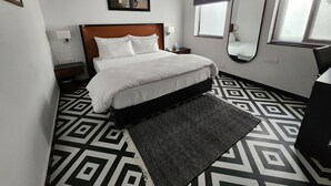 A hotel room with a bed, a nightstand, a bedside table, a mirror, a window with blinds, and a geometric patterned floor. - Stay Shaandaar (Amritsar)