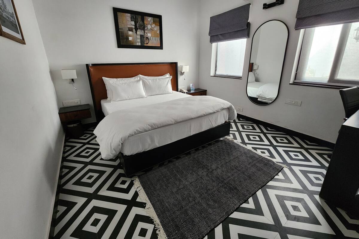 Habitación de hotel con cama, mesita de noche, espejo, ventana con persianas y suelo de estampado geométrico.