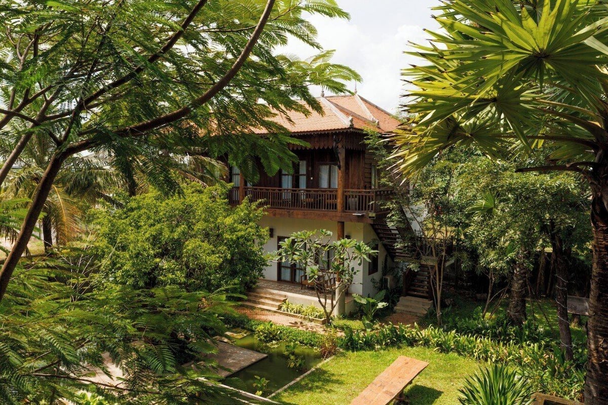 A traditional wooden house with a balcony, surrounded by lush greenery and a small pond.